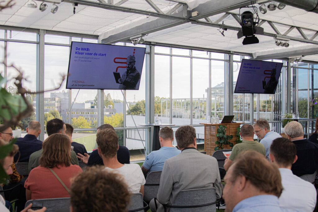 Een sfeerimpressie van het evenement WKB: klaar voor de start. De zaal vol mensen en twee schermen aan de voorkant van de zaal waar de presentatie op klaar staat.